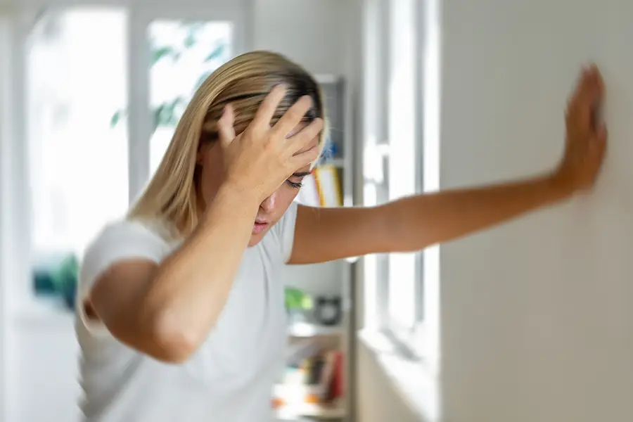 Woman steadying herself against a wall with one hand on her forehead, shown with a blurred effect to convey vertigo or dizziness.